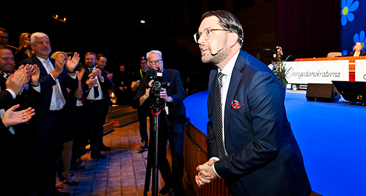 Jimmie Åkesson står vid en scen. Personer i publiken står upp och applåderar honom.