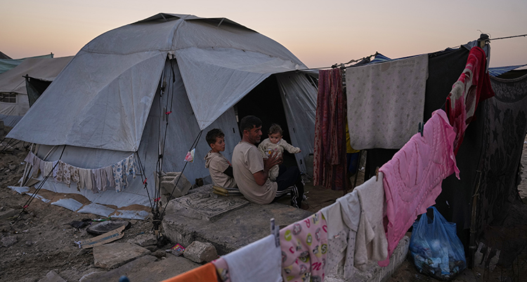 Han sitter med sina små barn utanför ett tält. Kläder hänger på tork.