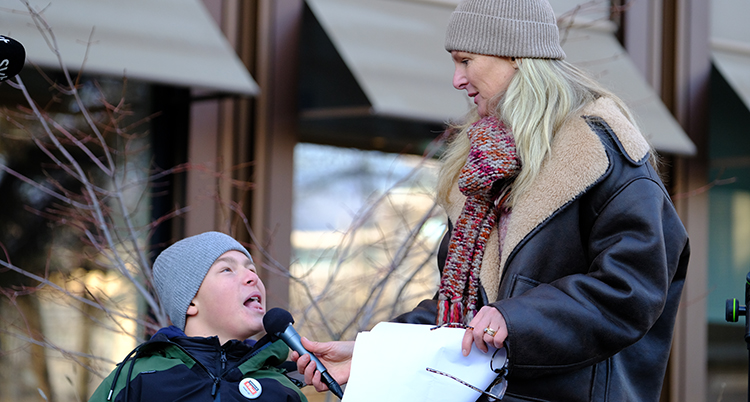 Hon håller i en mikrofon framför sin son som sitter ner i en rullstol.