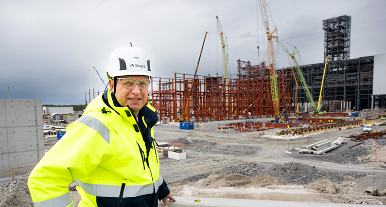 Han har en hjälm på sig. Han står framför ett stort bygge.