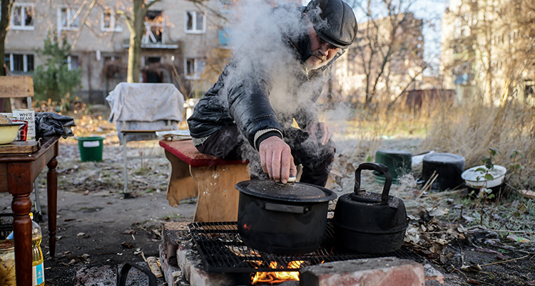 Han sitter framför en eld. Det röker från elden. Husen bakom är trasiga.