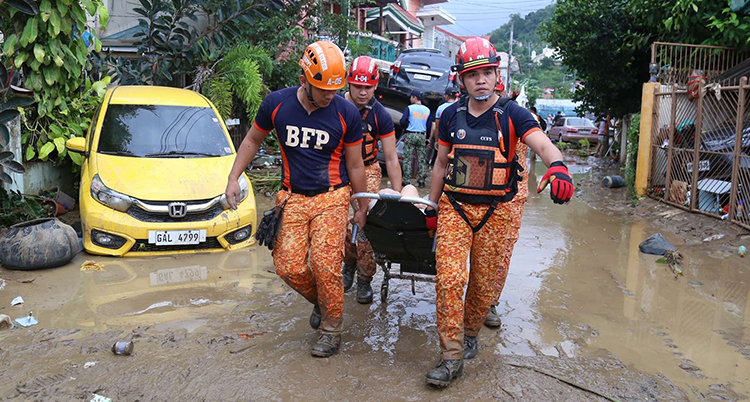 Flera personer i orange kläder bär på en bår. De går på en lerig väg.