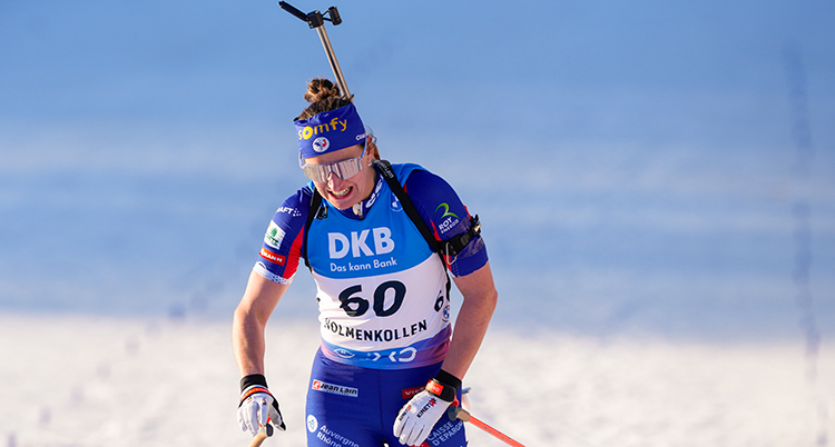 Hon åker skidor med ett gevär på ryggen. Hon har skidkläder och solglasögon.