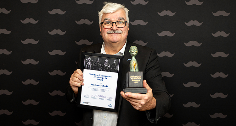 Han håller ett diplom och en staty i famnen. Han har glasögon och mustasch.