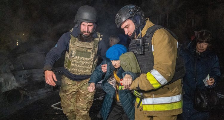 Två män från räddningstjänsten håller i en äldre kvinna. Det är mörkt ute.
