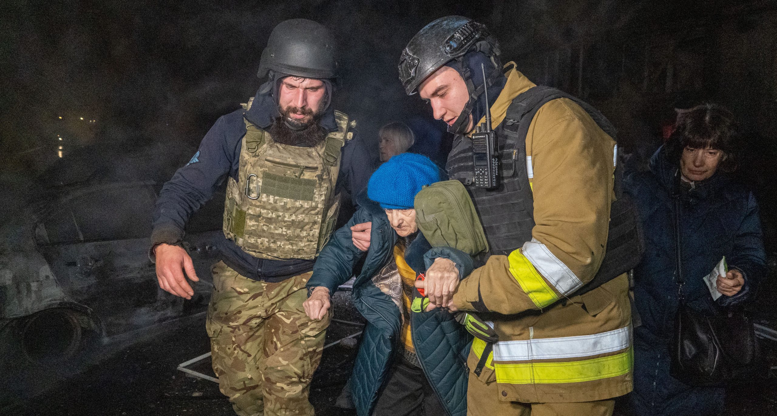 Två män från räddningstjänsten håller i en äldre kvinna. Det är mörkt ute.