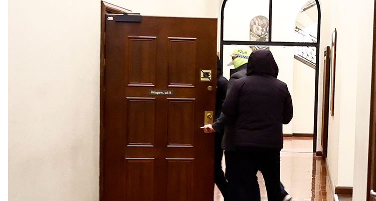 Mannen är bakom en en öppen dörr. Två poliser leder honom in genom dörren.