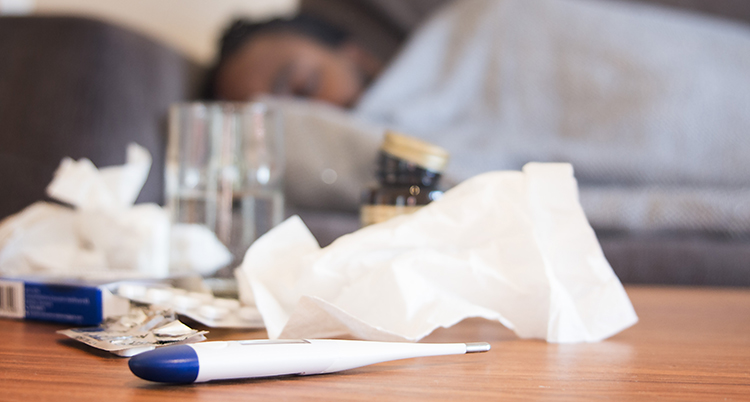 En person ligger i sin soffa. Det ligger papper och en termometer på ett bord.