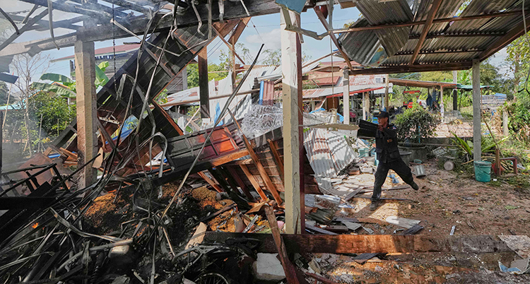 Ett hus är helt förstört. Det brinner. En person försöker släcka elden med vatten.