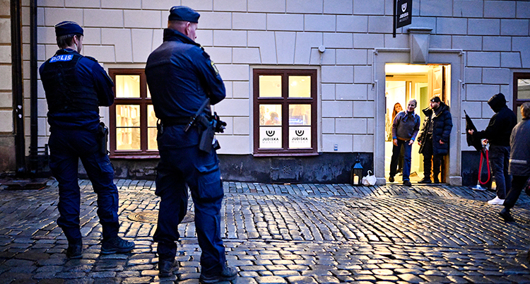 Två poliser står utanför ett hus i Gamla stan i Stockholm. I huset finns Judiska museet. Några personer tittar ut från museets dörr och tittar på poliserna.