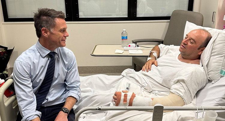 En man med slips sitter på sängen på ett sjukhus. En man ligger i sängen.