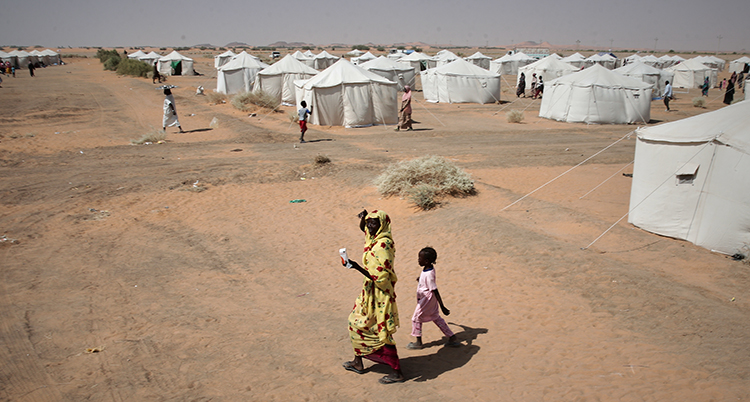De går i sand. Runt dem syns flera tält.