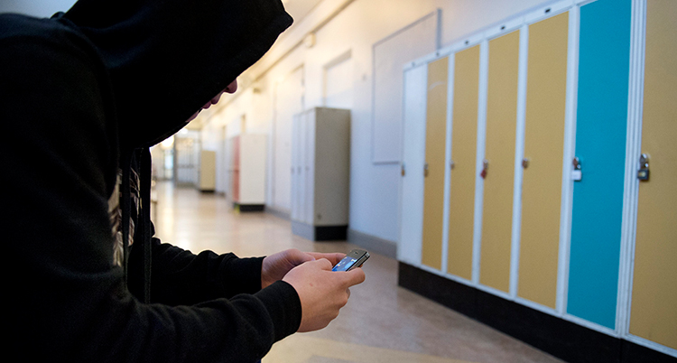 En ung pojke sitter med en mobil i en tom korridor med skåp i skola.