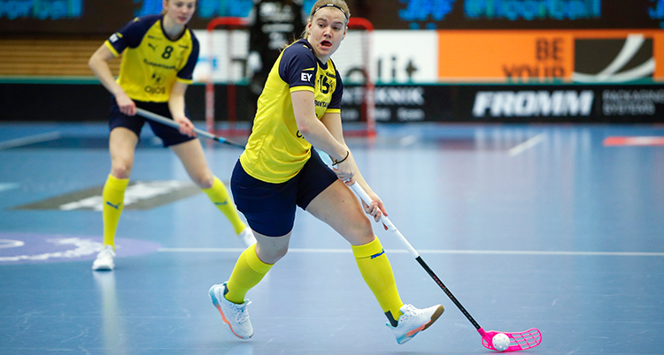 Hon spelar innebandy. Hon håller i en klubba och skjuter iväg bollen.