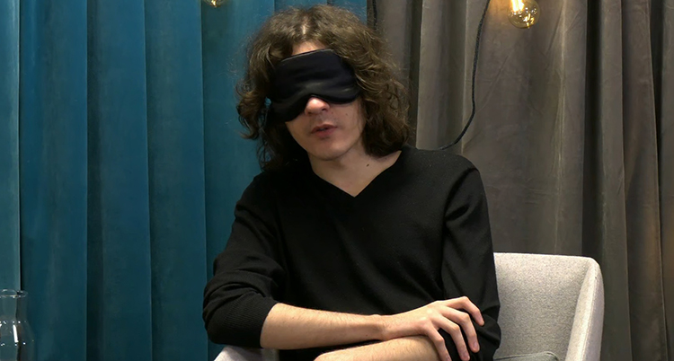 Han sitter på en stol. Han har en svart mask för ögonen.