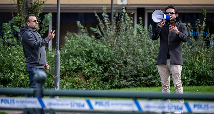 Två män står på en gata. En filmar, den andra pratar i en megafon.