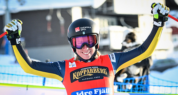 Hon är glad och sträcker ut armarna. Hon har skidglasögon på sig.