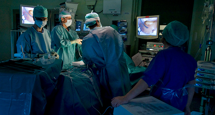 De står runt en säng i ett sjukhus och opererar. Det finns skärmar med bilder omkring dem.