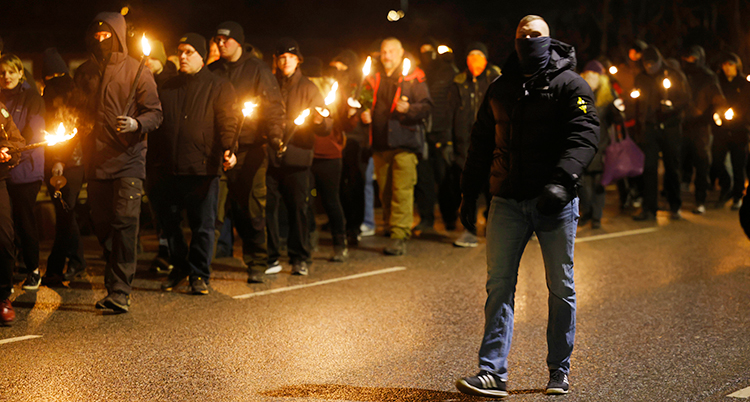 Det är mörkt ute. Människor går på en gata. De har facklor i händerna.