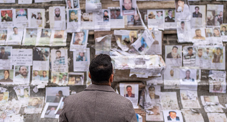 Mannan står med ryggen mot kameran. Väggen är fylld av papper och bilder på personer.