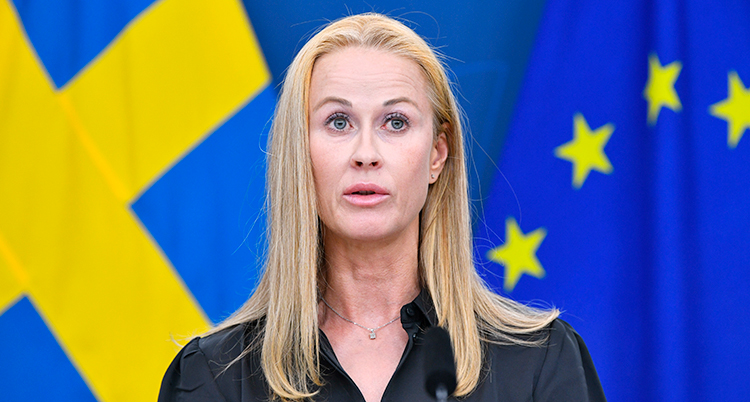 Hon har ljust långt hår. Hon står och pratar i en mikrofon. Bakom finns Sveriges flagga och EUs flagga.