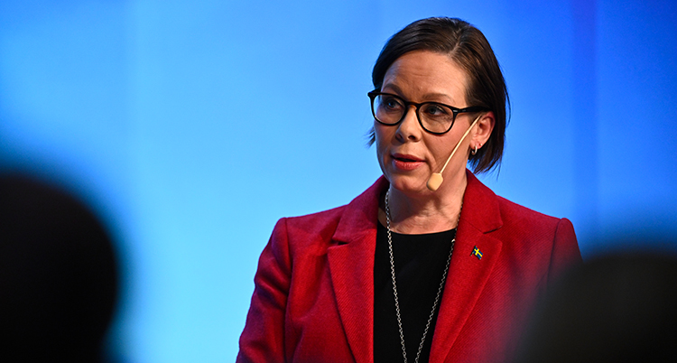 Hon har kavaj och en tröja under. Hon har en mikrofon vid munnen.