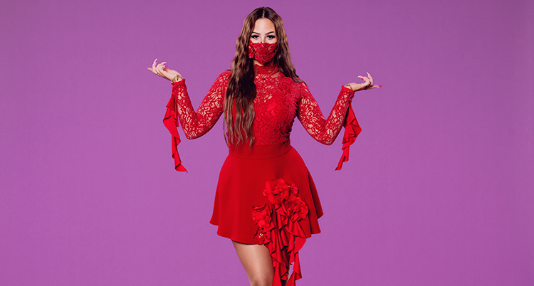 Hon har en röd klänning och långt mörkt hår. Hon tittar in i kameran. Hon har en mask över näsan och munnen.