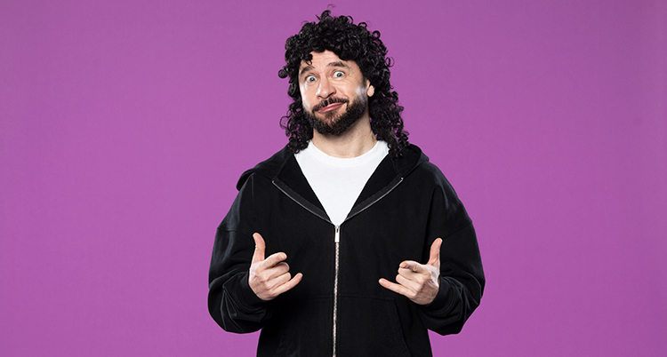 Han har på sig en mörk, lockig, peruk. Han ger en tokig min.