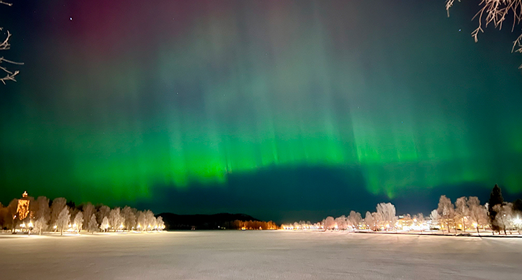 Det är mörkt ute. Det är snö på älven och träden. På himlen syns grönt norrsken.