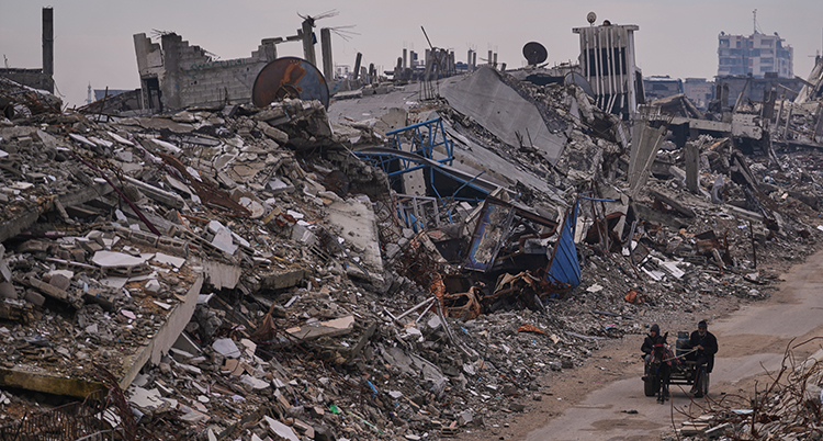 Två personer går på en torr väg. Det är förstörda hus intill.