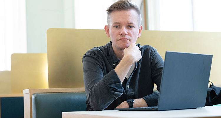 Han sitter vid ett bord med en dator framför sig. Han tittar in i kameran.