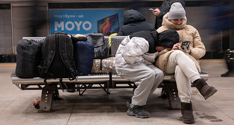 De sitter på en bänk och vilar och tittar i mobil. De har tjocka jackor.