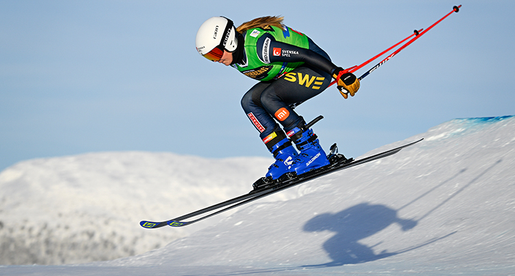 Hon hoppar med skidor och stavar. Det syns en skugga i snön under.
