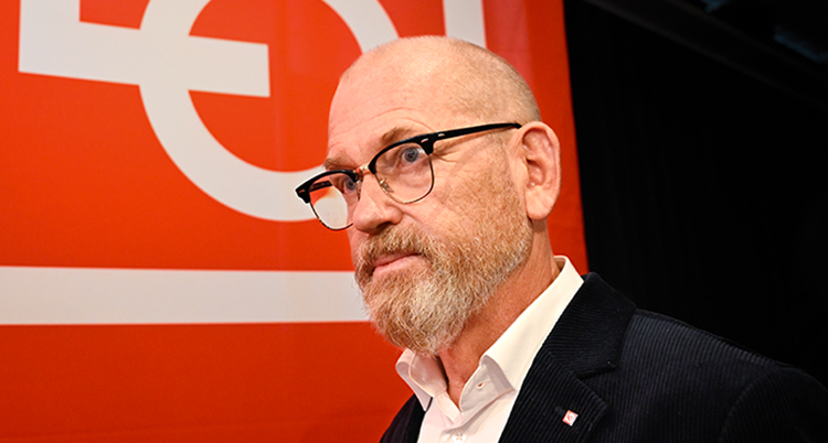 Han har skägg och glasögon. Han står framför en röd skylt för LO.