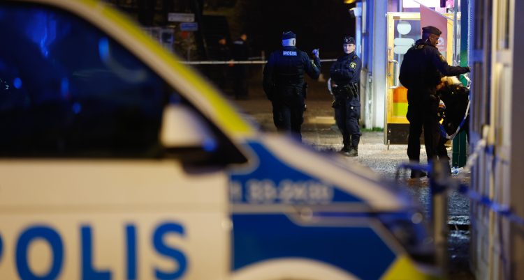 Några poliser står utanför en butik på kvällen. En polisbil står parkerad.