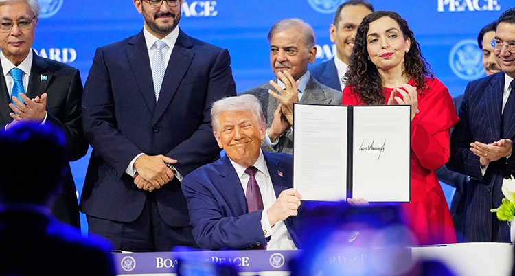 Han sitter vid ett bord med människor bakom sig. Han håller upp en uppslagen pärm med papper.