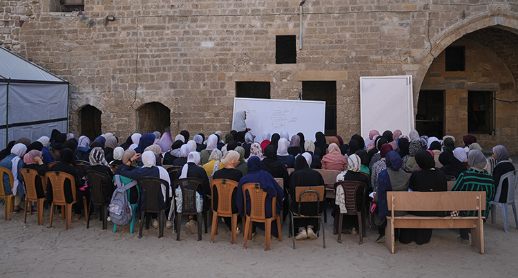 En stor grupp barn sitter på stolar utomhus. En läarare visar något på en vit tavla.