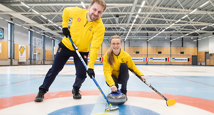 De är på en is för curling. Han håller i en sop för curling och är glad. Hon håller i en sten och en sop och är glad. De tittar in i kameran.