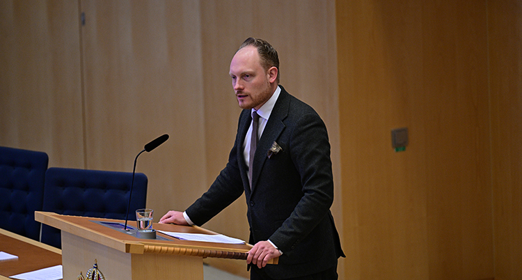 Han står framför en mikrofon i riksdagen.