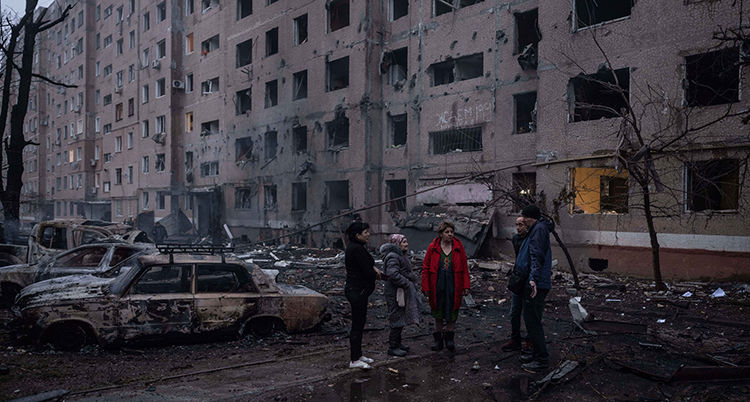 Människor står framför ett trasigt hus. En trasig bil står intill.