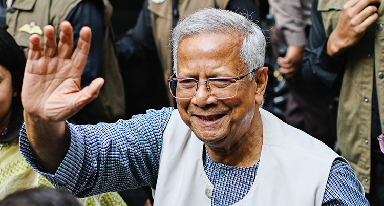 Han har grått hår och glasögon. Han vinkar med ena handen.