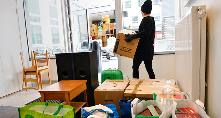 En person håller på att flytta och lyfter en kartong. På golvet står kartonger och påsar.