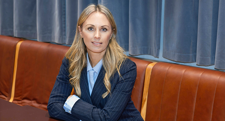 Hon sitter på en soffa med armarna i kors. Hon tittar in i kameran.