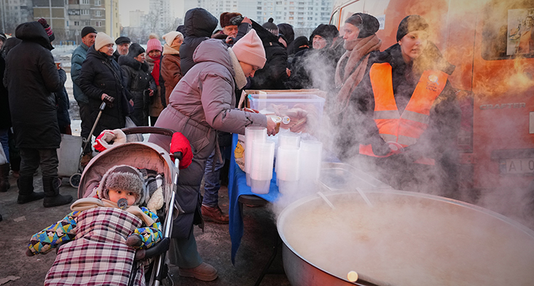 Många männsikor är samlade vid en stor rykande gryta utomhus. En kvinna med barnvagn häller upp soppa.