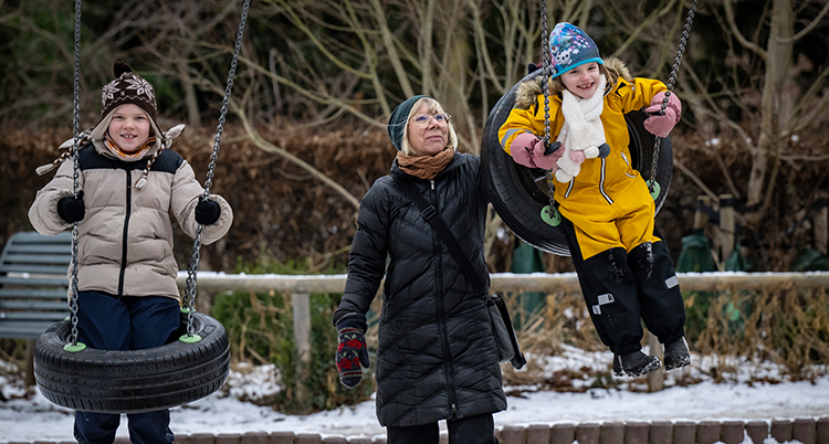 En flicka och en pojke sitter i varsin gunga. Deras farmor hjälper dem att få fart.