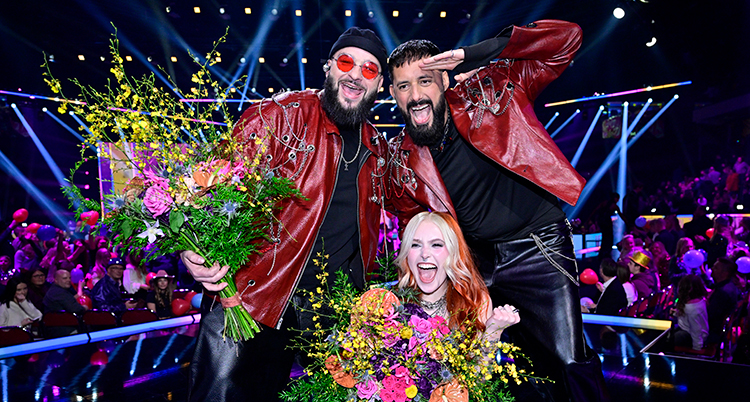 De firar segern på scen. De håller i varsin stor bukett blommor.