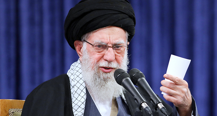 Han är 86 år. Han har vitt skägg och glasögon och en turban på huvudet. Han pratar i en mikrofon.