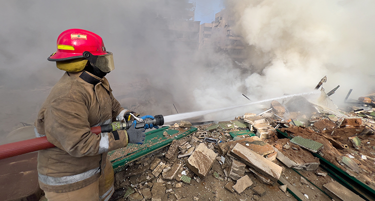 En brandman står och sprutar vatten på ett hus som är helt förstört. Det kommer rök från huset.
