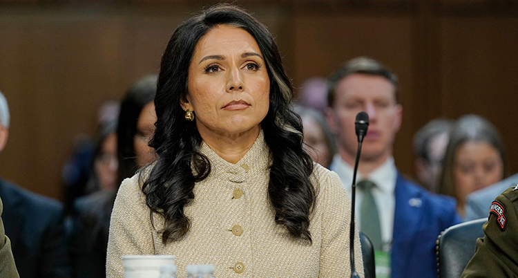 Hon ser allvarlig ut. Hon sitter i ett rum med många andra. Hon har en mikrofon framför sig.