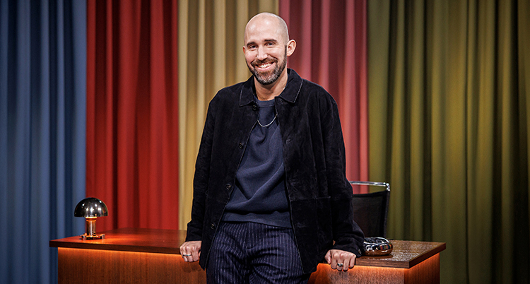 Han lutar sig mot ett skrivbord. Han ler och tittar in i kameran. Han har rakat huvud.